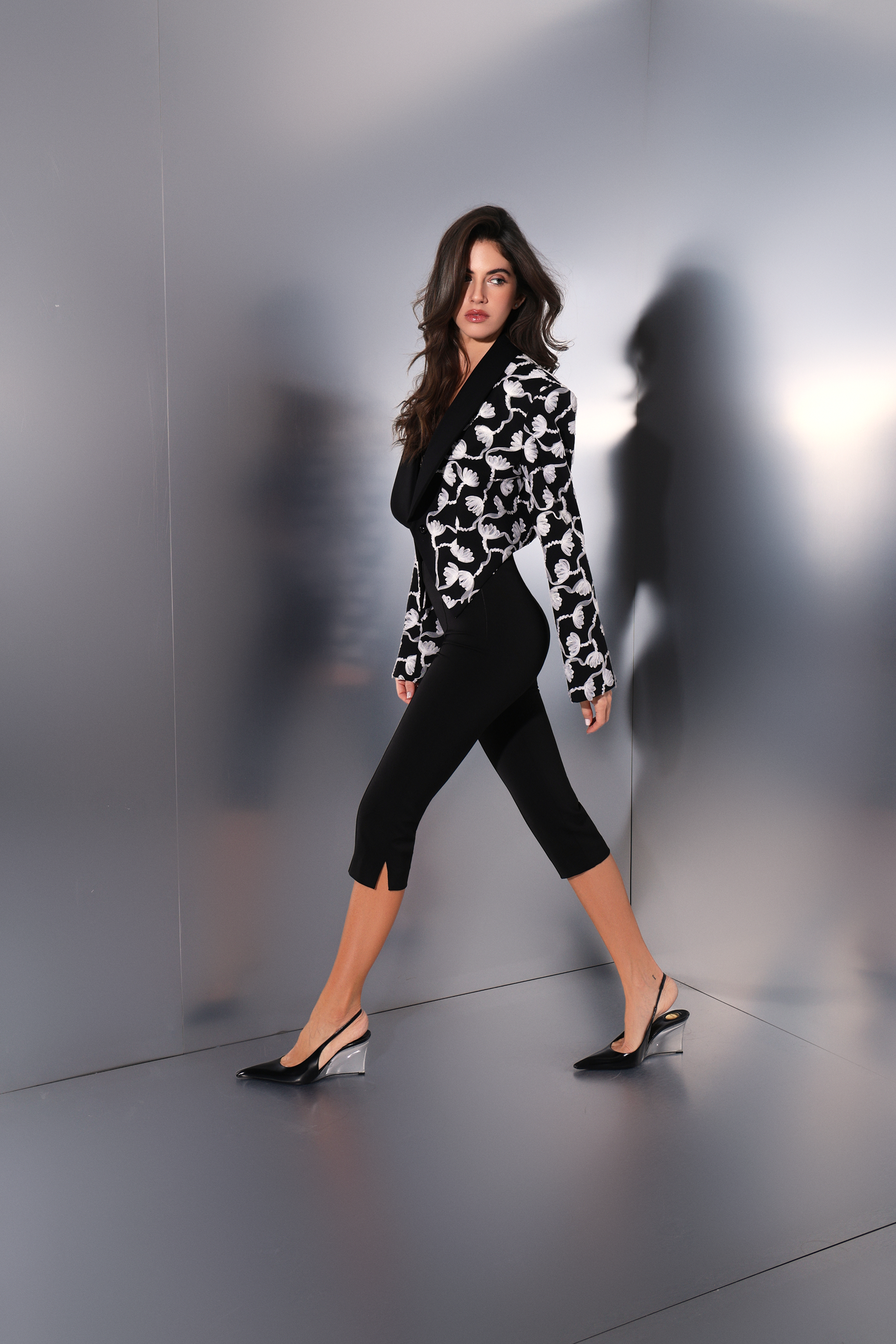 Woman in a black and white patterned top and black pants standing against a reflective gray wall.