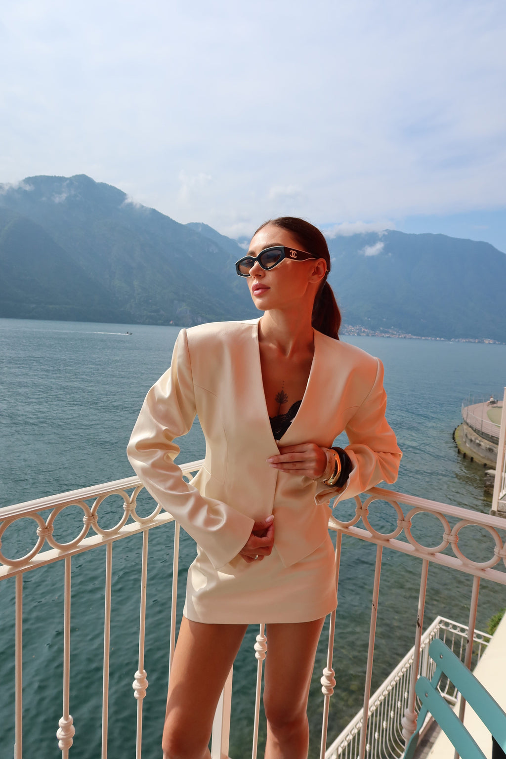 Woman in a white suit standing on a balcony overlooking a scenic view of water and mountains.