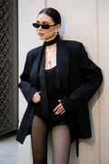 Person wearing a black outfit with a coat and high heels, standing against a neutral wall.