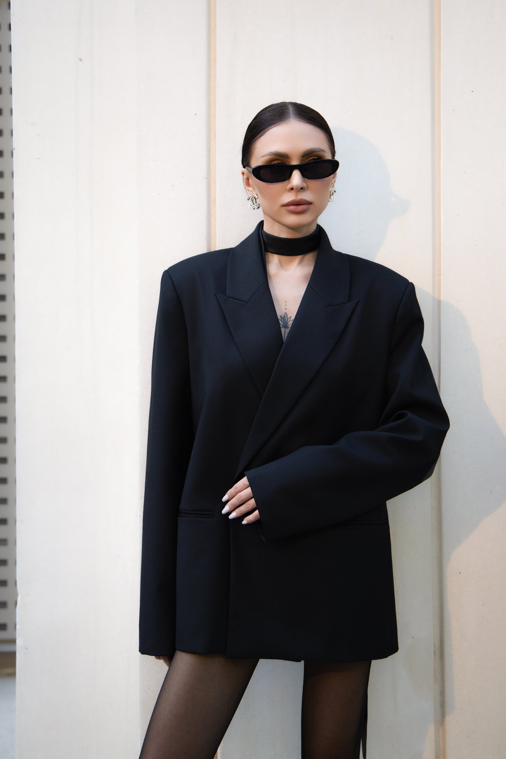 Person wearing a black coat and sunglasses against a light-colored wall.